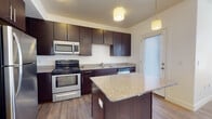 This single-floor residence offers a thoughtfully designed layout combining functionality and modern finishes throughout. The kitchen serves as a central highlight, showcasing rich dark-espresso cabinetry with sleek bar-pull hardware, speckled granite countertops, and a full suite of stainless steel appliances including a refrigerator, range, microwave, and dishwasher. A central island with a matching granite surface provides additional prep space and casual seating. Warm wood-look flooring runs throughout the kitchen and living areas, while pendant lighting adds an inviting ambiance. A door with sidelight window in the kitchen area allows natural light to filter in. The bathroom is finished consistently with the kitchen, featuring the same dark cabinetry, granite vanity top, a large mirror, and a full tub-shower combination with clean white surroundings and tile flooring. Bedrooms are carpeted and bright, benefiting from natural light through window blinds. A dedicated laundry area houses a full-sized washer and dryer alongside the water heater, adding everyday convenience. Overall, the space presents a cohesive, modern aesthetic with practical amenities throughout.