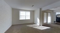 This single-floor space offers a clean, straightforward layout designed for comfortable everyday living. The main living area is finished with wall-to-wall carpet flooring in a neutral tone, providing a warm and practical foundation throughout the space. Crisp white walls keep the interior feeling open and airy, offering a versatile backdrop that adapts easily to a variety of furnishing styles.

Natural light is a defining feature of this space. A large multi-pane window fitted with horizontal blinds serves as the focal point of the main room, allowing generous sunlight to stream in and cast bright, geometric patterns across the floor. A secondary window beside the entry door further enhances the brightness, ensuring the space remains well-lit throughout the day.

The layout includes a clearly defined entryway with a door featuring a built-in window with blinds, offering both natural light and a practical point of access. Structural column details near the entry add a subtle architectural element to the otherwise clean interior. The ceiling is smooth and white, with a centrally located HVAC vent indicating a climate-controlled environment. Overall, the space presents a neat, move-in ready interior with a light-filled, neutral aesthetic.