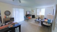 This single-floor living space is designed with comfort and functionality in mind. The living room serves as the central hub, bathed in abundant natural light streaming through large vertical-blind-covered windows and a sliding glass door that spans much of one wall, offering an airy, open feel. A ceiling fan is centrally positioned overhead, contributing to the room's ventilation and comfort.

The flooring throughout is soft, neutral-toned carpeting, which adds warmth underfoot. A fireplace with a clean, white surround serves as a notable architectural focal point along the main wall, lending the space a cozy character. The walls are painted in a light, neutral tone that amplifies the brightness of the room.

The layout accommodates distinct functional zones within the open floor plan. A seating area is anchored by a low-profile coffee table atop a plush area rug, surrounded by a sofa and an accent chair. A media console holds a flat-screen television, accompanied by decorative artwork above. A separate dining area is visible near the entrance, featuring a dark-toned table. Overall, the space presents a welcoming, well-organized atmosphere with versatile furnishing arrangements.