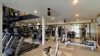 This two-floor space includes a dedicated fitness center that is thoughtfully designed to support a comprehensive workout experience. The gym area is generously sized, featuring an open layout with a patterned floor that adds a modern aesthetic touch. Recessed lighting throughout the ceiling ensures bright, even illumination across the entire space, while multiple ceiling fans promote comfortable air circulation during workouts.

The fitness center is equipped with a wide array of cardio machines, including treadmills, stationary bikes, and rowing machines, all positioned to allow ample space between equipment. A strength training section features a cable machine system and a full dumbbell rack, catering to varied fitness routines. Large mirrors line the walls, enhancing the sense of space while allowing users to monitor their form. Mounted flat-screen televisions are also visible, providing entertainment during exercise sessions. Natural light filters in through windows along one side of the room, contributing to an inviting and energizing atmosphere. The overall design is clean, functional, and well-maintained, making it a practical and appealing fitness facility.