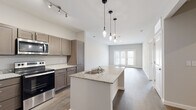 This single-story residence in Little Rock, Arkansas, offers a thoughtfully designed open-concept layout with contemporary finishes throughout. The kitchen serves as a central hub of the space, anchored by a large granite-topped island with a double-basin sink and stylish pendant lighting overhead. Taupe shaker-style cabinetry provides ample storage, complemented by granite countertops along the perimeter and a full suite of stainless steel appliances, including a refrigerator, electric range, and microwave. Wood-style plank flooring extends seamlessly from the kitchen into the adjacent living area, which is bathed in natural light from a set of glass French doors that open to the exterior. The open dining and living zones offer generous space with a neutral color palette of soft grays and warm whites, creating a clean, inviting atmosphere. The bedrooms feature the same durable wood-style flooring, ceiling fans, and windows that allow natural light in. The bathroom is finished with wood-style flooring, neutral toned walls, and a full tub and shower surround. Recessed lighting and track lighting add to the well-lit, modern character of this single-floor space.