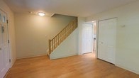 This two-floor home offers a bright and practical layout designed for comfortable everyday living. The main level showcases light-toned hardwood flooring that extends throughout the open area, lending warmth and continuity to the space. Soft sage-green walls provide a calm, neutral backdrop that complements the natural wood tones found throughout.

A central staircase with a light wood banister and classic spindle railing serves as a functional focal point, connecting the main floor to the upper level. The staircase is neatly integrated into the layout, with clean white trim and baseboards framing the transition between floors.

Adjacent to the staircase, a set of white six-panel doors provides access to additional rooms, with a glimpse into what appears to be a kitchen area visible through an open doorway. The flush-mounted ceiling light fixture provides even, ambient illumination across the lower level.

The overall design is clean and understated, with consistent finishes throughout — white doors and trim, neutral wall tones, and warm flooring — creating a cohesive and move-in-ready environment. The space is well-maintained and presents a functional, organized flow between areas, making it a practical and welcoming home across both floors.