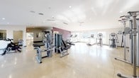 The Main Fitness Center is a spacious, single-floor facility designed to support a wide range of workout routines. The interior features polished concrete floors throughout, recessed lighting, and high ceilings that contribute to an open, airy atmosphere. Large windows along one side of the facility allow ample natural light to flood the space, offering views of the outdoors and creating an energizing environment for exercise.

The cardio zone is well-appointed with a row of Matrix treadmills, an elliptical trainer, a Matrix stair climber, and multiple spin bikes — all positioned near the windows to take advantage of natural light. The strength training area is equipped with a variety of HOIST and Matrix cable machines, selectorized weight machines, and a dumbbell rack with a wall mirror, providing options for both isolated and compound movements.

A bold burgundy accent wall displays motivational text, adding character and energy to the workout environment. A second green-lettered motivational quote adds to the facility's inspiring aesthetic. A dedicated Game Zone room with a glass door is accessible from the main floor. An informational bulletin board near the entrance keeps members up to date on community resources. The facility is clean, well-maintained, and thoughtfully laid out to maximize usability for all fitness levels.