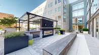 Located at 517 N Shipley St in downtown Wilmington, Delaware, this contemporary outdoor amenity level showcases a thoughtfully designed urban retreat. The space is anchored by a sparkling swimming pool equipped with stainless steel railings and accessibility lifts, surrounded by a generous pool deck lined with sleek lounge chairs and lush greenery. Raised planters filled with manicured shrubs and shade trees create natural divisions throughout the outdoor areas, lending a sense of privacy and tranquility amid the urban setting. A dedicated lounge terrace features modern rounded white chairs arranged around a small side table, ideal for casual gatherings. A separate wood-decked terrace offers relaxed seating with Adirondack chairs and a cornhole board, providing a recreational retreat. Paved walkways meander through curated garden beds, connecting the various zones of the amenity level seamlessly. The exterior façade is defined by a mix of dark brick and light-toned panel cladding, complemented by expansive glazing that allows natural light to interact with the surrounding spaces. The main entrance is marked by a set of sleek double glass doors framed in dark steel, reinforcing the building's modern architectural character. The overall ambiance balances urban sophistication with approachable outdoor living.