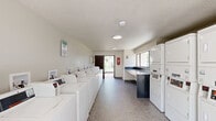 Located at 15690 Bull Run Rd in Miami Lakes, Florida, this communal laundry facility is a practical, single-floor space designed to efficiently serve its users. The room is organized into two distinct machine zones: a row of commercial-grade top-loading washers lines one side, while a bank of stacked washer-dryer units runs along the opposite wall, maximizing the available floor area. Each machine is coin-operated, providing straightforward usability for residents. A built-in dark countertop folding station is conveniently positioned near a window, offering natural light and a dedicated surface for sorting and folding laundry. The flooring throughout is a speckled terrazzo tile, lending the space a clean, durable, and easy-to-maintain finish. The walls are finished in a neutral off-white with a textured ceiling, creating a bright and functional atmosphere. A prominent entrance features a dark slatted louvered door that opens outward to a lush, landscaped outdoor area with mature trees and a brick-paved walkway, allowing natural light and fresh air to flow through. Safety is also addressed with a clearly visible fire extinguisher mounted near the entrance. Posted signage on the walls communicates laundry room rules and hours for users.