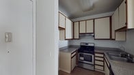 This single-floor space is designed with practicality and functionality in mind. The kitchen serves as a central feature, showcasing a stainless steel gas range paired with an integrated range hood overhead, providing efficient ventilation during cooking. Granite-style countertops offer durable workspace on either side of the range, while an abundance of upper and lower cabinetry — finished in a cream tone with warm brown wood trim accents — delivers generous storage capacity. The cabinetry spans multiple walls, creating a well-organized and efficient layout that maximizes every inch of available space. The flooring transitions to hardwood-style planks, adding warmth and continuity throughout. Neutral-toned walls and simple, clean finishes contribute to a bright, understated ambiance. The layout is compact yet thoughtfully arranged, making the most of the available footprint. The space presents a straightforward, move-in-ready environment suited for everyday living, with classic design elements that offer a solid foundation for personalization.