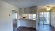 This single-floor residential unit offers a practical and functional layout designed to maximize the use of available space. At the heart of the home is an open-concept kitchen, thoughtfully designed with a peninsula-style pass-through counter that connects the cooking area to the main living space. The kitchen is equipped with white appliances, upper cabinetry, and an additional set of open built-in shelves overhead, providing ample storage and display space.

The flooring transitions from rich, dark hardwood in the main living and kitchen areas to plush carpeting in the adjoining bedroom, offering both aesthetic contrast and comfort underfoot. A textured popcorn ceiling runs throughout, adding a classic residential character to the space.

Natural light is a notable feature, with windows visible from multiple vantage points, including a sliding glass door dressed with vertical blinds that leads to what appears to be an outdoor balcony or patio. The neutral color palette of warm whites and soft beiges throughout the unit creates a clean, understated ambiance. The open sightlines between the kitchen, living area, and bedroom contribute to an airy, connected feel throughout the home.
