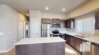 Located at 12501W Sixth Ave in Airway Heights, Washington, this single-floor residence offers a thoughtfully designed layout with a clean, modern aesthetic throughout. The kitchen serves as a functional centerpiece, showcasing rich dark wood cabinetry, crisp white countertops, a central island, and a full suite of stainless steel appliances including a refrigerator, electric range, built-in microwave, and dishwasher. Recessed lighting and light hardwood-style flooring complete the kitchen's contemporary feel. The open living area extends from the kitchen, featuring the same light hardwood flooring and a wall-mounted mini-split system for climate control. A sliding glass door floods the space with natural light and provides views of the surrounding neighborhood. The bedrooms are finished with plush carpeting and neutral-toned walls, with multiple windows allowing ample daylight. The bathroom features a white vanity with an integrated sink, a large mirror, and a walk-in shower, all finished in a clean, neutral palette. The overall ambiance of the residence is bright, modern, and move-in ready, with consistent finishes throughout that create a cohesive and comfortable living environment.
