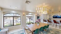Located at 1111 North Mission Park Boulevard in Chandler, Arizona, this single-floor space offers a seamlessly connected living room, dining room, and kitchen within a well-appointed footprint. The living room is a standout, boasting dramatic floor-to-ceiling windows that frame lush outdoor views of mature trees and landscaped grounds, flooding the interior with natural light. The soaring ceiling height amplifies the sense of openness, while warm gold sconce lighting adds an inviting ambiance. The dining area continues the refined aesthetic with arched windows framed in dark trim, a large wood communal table, eclectic seating, and a statement capiz shell chandelier with gold detailing. A vibrant area rug and curated wall art complete the look. The kitchen features granite countertops, modern wood cabinetry, a built-in wine cooler, and a striking stacked stone accent wall with a mounted flat-screen television. Large-format tile flooring runs throughout, tying the spaces together cohesively. The overall design blends Mediterranean-inspired architectural elements with contemporary furnishings, creating a bright, welcoming, and highly functional space.
