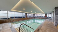This amenity floor at 2121 Avenue Saint-Mathieu in Montréal presents a thoughtfully designed wellness facility combining an indoor pool area and a fitness centre. The indoor pool is the centrepiece of the space, finished with aqua mosaic tile coping and surrounded by large-format stone-toned floor tiles. A dramatic coffered ceiling with integrated warm LED cove lighting runs the perimeter, creating a calm, resort-like ambiance. Floor-to-ceiling windows wrap around the pool on multiple sides, flooding the area with natural light and offering sweeping panoramic views of the Montréal skyline and Mont-Royal. The pool deck includes entry handrails, a safety ring station, and a stretcher board, reflecting a well-maintained, safety-conscious environment. Adjacent to the pool, textured grey stone-clad walls enclose change room and sauna access points, adding a contemporary spa aesthetic. The connected fitness centre features rubber flooring, a mirrored free-weight alcove with a full dumbbell rack and bench, stability balls, cardio equipment, and wood slat ceiling accents. Together, these amenities form a cohesive, upscale wellness environment designed for everyday use.