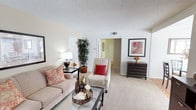 This single-floor residence offers a comfortable and thoughtfully designed living environment with a warm, neutral aesthetic throughout. The living room serves as the centerpiece of the space, featuring wall-to-wall carpeting in a soft beige tone that contributes to the room's inviting atmosphere. Light-colored walls paired with a textured ceiling enhance the sense of openness, while a central HVAC vent ensures year-round comfort.

The living area is generously proportioned, providing ample room for a full sofa ensemble, accent chairs, and a glass-top coffee table, all arranged to encourage a natural flow and comfortable conversation. A dark wood dresser-style accent cabinet adds a rich contrast to the lighter surroundings, complemented by framed wall art that lends a polished, curated feel to the space.

A tall indoor plant positioned near the entryway corridor introduces a natural element that softens the room and adds visual height. The layout opens conveniently through a doorway into a hallway connecting to additional areas of the residence, suggesting a functional and accessible floor plan. Overall, the space balances practicality with a clean, welcoming aesthetic suitable for everyday living.
