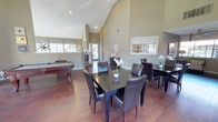 This single-floor community clubhouse offers a well-appointed and versatile space designed to accommodate a variety of social and professional activities. The main gathering area is immediately impressive, featuring dramatic vaulted ceilings that soar overhead and create a grand sense of openness and volume. Rich hardwood flooring runs throughout, adding warmth and a polished aesthetic to the space.

The open-concept layout seamlessly integrates a dining area, furnished with a dark wood table and leather chairs, alongside a dedicated recreation zone that includes a full-size billiards table. Large windows fitted with striped Roman shades allow natural light to filter in, brightening the warm neutral tones of the walls. A decorative console and thoughtful accent pieces contribute to the refined yet welcoming ambiance.

A separate business center provides a quieter, more focused environment. This room features a row of computer workstations along a dedicated desk surface, complemented by a comfortable lounge seating area with armchairs and a coffee table. Multiple windows line the walls, flooding the room with natural light and offering views of the surrounding exterior. Carpeted flooring and angled ceilings lend a relaxed, professional feel to this functional workspace, making it well-suited for residents seeking a productive environment outside the main gathering hall.