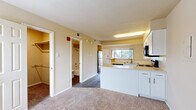 This efficiently designed single-floor unit offers a thoughtful open-concept layout that maximizes every square foot. The living room serves as the heart of the space, featuring warm neutral-toned carpet, high vaulted ceilings that lend a sense of openness, and a sliding glass door that opens onto a private balcony with outdoor views of surrounding greenery — a standout feature that fills the area with natural light.

The kitchen is positioned adjacent to the living area, separated by a peninsula-style breakfast bar with light-toned countertops. White cabinetry lines the walls, providing ample storage, while black appliances — including a refrigerator, electric range, built-in microwave, and dishwasher — offer a clean contrast. Dark hardwood-style flooring grounds the kitchen and entry areas.

The bathroom is clean and functional, featuring a full tub-and-shower combo with white tile surround, alongside overhead cabinetry for additional storage. Two closets provide practical storage solutions throughout the unit.

The overall ambiance is bright and neutral, with warm beige walls, a combination of carpet and hard flooring, and a simple, functional aesthetic throughout. This unit is well-suited for those seeking a streamlined, low-maintenance living space.