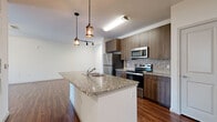This 1-bedroom, 1-bathroom unit is thoughtfully designed to maximize functionality and comfort across a single floor. The open-concept kitchen and living area forms the heart of the space, anchored by a central island with granite countertops and a built-in sink, illuminated by stylish pendant lighting. The kitchen is equipped with stainless steel appliances, including a refrigerator, range, and microwave, complemented by rich wood-toned cabinetry and a mosaic tile backsplash. Rich wood-look flooring flows throughout the main living areas, adding warmth and continuity. The spacious living room is bathed in natural light from large sliding glass doors that open to a private balcony, offering outdoor views and a ceiling fan for comfort. The bedroom provides a cozy retreat with plush carpeting, a ceiling fan, and windows that bring in additional natural light. A dedicated closet offers convenient storage. The bathroom is elegantly appointed with a dual-sink granite vanity, wood-toned cabinetry, a soaking tub with tile surround, and large-format tile flooring. A separate closet room provides additional organizational space, rounding out this efficiently designed and well-finished unit.