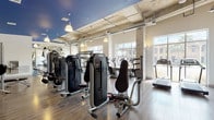 This well-appointed fitness center occupies a single open floor, designed to accommodate a comprehensive range of workout activities. The space is defined by soaring ceilings with exposed ductwork and structural elements, lending it an industrial character that is balanced by clean white walls and warm wood-look flooring throughout.

A striking navy blue ceiling accent delineates one section of the facility, adding a bold visual contrast to the otherwise bright, airy environment. Pendant lighting is suspended throughout, complementing the abundant natural light that pours in through large floor-to-ceiling windows lining the perimeter. These windows frame outdoor views of the surrounding landscape, including palm trees and neighboring architecture, enhancing the open atmosphere during workouts.

Textured stone-clad columns punctuate the interior, adding an architectural dimension to the space. The cardio zone features a row of treadmills and elliptical machines positioned directly in front of the windows, allowing users to enjoy the outdoor scenery. The strength training area is equipped with a comprehensive selection of Monolith cable and resistance machines arranged in organized rows. A free weights station includes a full dumbbell rack, barbells, and wall-mounted televisions for entertainment. Full-length mirrors expand the sense of space and support form monitoring during exercise.