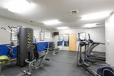 This fitness center offers a comprehensive workout environment thoughtfully arranged across a single level. The space is finished with durable rubber flooring throughout, providing a safe and functional surface ideal for a variety of exercise activities. Walls are painted in a two-tone scheme of gray and bold blue, lending the space an energetic and motivating atmosphere, while a textured ceiling with recessed fluorescent lighting ensures the entire area is brightly and evenly illuminated.

The main workout area features a well-stocked dumbbell rack with a full range of free weights, a flat workout bench, and a wall-mounted mirror that enhances the sense of space and allows for form checks during exercise. A heart rate target chart and exercise reference poster are displayed on the wall for user guidance, alongside a wall clock and a wall-mounted television. A water cooler station is conveniently positioned nearby for hydration.

A dedicated cardio alcove houses multiple treadmills, including a SportsArt model, providing ample space for cardiovascular training. The adjoining area accommodates a multi-station cable strength machine along with additional cardio equipment such as an elliptical or cycle trainer. A small seating area with chairs near the entrance offers a space to rest, and coat hooks line the wall for added convenience. The overall layout is practical, organized, and well-suited for a full-body workout routine.
