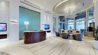 This single-floor lobby space makes a striking architectural statement through its thoughtful design and high-end finishes. At its centerpiece sits a curved, dark wood reception desk adorned with decorative carved detailing, positioned atop a patterned terrazzo floor with inlaid metallic accents that guide movement through the space. Behind the desk, a large-scale teal mosaic tile feature wall commands attention, providing a bold and vibrant focal point that anchors the entire room.

The ceiling design is equally impressive, featuring a layered circular coffered detail from which a dramatic multi-tiered crystal chandelier descends, adding glamour and reflecting light throughout the space. Recessed and accent lighting complement the natural light that filters in through floor-to-ceiling windows along the far wall.

A comfortable lounge seating area occupies one side of the lobby, furnished with modern curved gray chairs and a cream sofa accented with red cushions, arranged around sleek glass-topped side tables. The overall aesthetic blends contemporary design with luxurious materials, creating a polished, professional ambiance suited to an upscale hospitality or commercial environment.