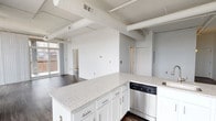 This contemporary urban loft showcases a thoughtfully designed open-concept layout that seamlessly integrates the living, dining, and kitchen areas into one expansive, light-filled space. The industrial character of the unit is defined by exposed ductwork and structural beams along the ceilings, adding architectural interest and an authentic loft aesthetic. Rich, dark-toned hardwood flooring runs throughout the main living areas, providing a striking contrast against the crisp white walls and ceilings.

The kitchen is well-appointed with white shaker cabinetry, granite-speckled countertops, a peninsula island with a sink, and a full suite of stainless steel appliances including a dishwasher, range, and built-in microwave. The open kitchen flows naturally into the generous living area, making the space ideal for entertaining.

Floor-to-ceiling windows along multiple walls bathe the interior in abundant natural light and offer sweeping urban cityscape views. A sliding glass door provides access to a private balcony, further extending the connection to the surrounding city.

The bathrooms feature granite-topped vanities with white cabinetry, classic white tile flooring, and clean, modern fixtures. The overall ambiance of the space is bright, airy, and urban — a polished blend of industrial character and contemporary comfort.