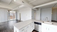 This contemporary urban loft showcases a thoughtfully designed open-concept layout that seamlessly integrates the living, dining, and kitchen areas into one expansive, light-filled space. The industrial character of the unit is defined by exposed ductwork and structural beams along the ceilings, adding architectural interest and an authentic loft aesthetic. Rich, dark-toned hardwood flooring runs throughout the main living areas, providing a striking contrast against the crisp white walls and ceilings.

The kitchen is well-appointed with white shaker cabinetry, granite-speckled countertops, a peninsula island with a sink, and a full suite of stainless steel appliances including a dishwasher, range, and built-in microwave. The open kitchen flows naturally into the generous living area, making the space ideal for entertaining.

Floor-to-ceiling windows along multiple walls bathe the interior in abundant natural light and offer sweeping urban cityscape views. A sliding glass door provides access to a private balcony, further extending the connection to the surrounding city.

The bathrooms feature granite-topped vanities with white cabinetry, classic white tile flooring, and clean, modern fixtures. The overall ambiance of the space is bright, airy, and urban — a polished blend of industrial character and contemporary comfort.