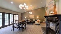 This single-floor space offers a thoughtfully designed open-plan layout that seamlessly combines a dining area and a comfortable lounge. The interior is finished in warm, neutral earth tones — taupe walls paired with light carpeting — creating an inviting and cohesive ambiance throughout.

Two elegant chandelier light fixtures hang from the vaulted ceiling, providing a refined focal point while recessed lighting supplements the overall illumination. The generously proportioned ceiling height enhances the sense of openness and airiness within the space.

The dining area, positioned near French doors that allow natural light to filter in, features a dark wood table surrounded by matching cross-back chairs, offering a practical yet stylish setting. Adjacent to this, the lounge area is furnished with a dark sofa and patterned accent chairs arranged around a central coffee table, creating a relaxed and social atmosphere.

A fireplace with a dark surround anchors one end of the room, adding a warm, welcoming element to the space. Decorative touches such as framed artwork, table lamps, and accent vases contribute to the polished, comfortable character of this well-appointed interior.