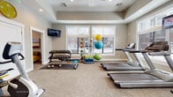 This fitness center is designed to support a comprehensive workout experience within a bright, welcoming environment. The main exercise area is spaciously laid out with neutral-toned carpet flooring and benefits from abundant natural light streaming through large windows that overlook the surrounding exterior. A tray ceiling with recessed lighting and a ceiling fan adds architectural definition while maintaining an airy atmosphere.

The cardio zone is well-appointed with multiple treadmills, elliptical cross-trainers, and stationary recumbent and upright bikes, several of which feature individual mounted screens for entertainment during workouts. A dedicated strength training section includes a multi-station cable machine, a full rack of dumbbells ranging across various weights, a flat workout bench, and an assortment of medicine balls, exercise balls, and foam rollers stored on an organized rack. Wall-mounted mirrors in the strength area enhance the sense of space and allow users to monitor their form. A wall-mounted television is also present for additional viewing.

Adjacent to the main fitness floor is a compact children's play nook, thoughtfully separated to create a family-friendly environment. This cozy space features tall windows, a wall-mounted TV, a low activity table, toy storage bins, and a vibrant yellow sectional sofa with bean bag seating, providing a comfortable area for younger visitors.