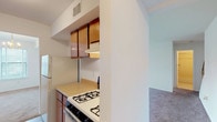 This two-story residence at Durham Woods, Unit 2N, offers a straightforward and functional layout spread across two levels, connected by a carpeted staircase with a wood-railed banister. The main level features a generously sized living area finished with wall-to-wall carpeting and a sliding glass door that opens to a balcony, allowing natural light and outdoor views to filter into the space. A staircase with a skylight above introduces additional natural light to the lower level. Adjacent to the living area, the kitchen is laid out in a galley style with warm-toned wood cabinetry lining both walls, light laminate countertops, a stainless steel sink, a gas range, and tile flooring. A louvered closet door within the kitchen provides convenient storage access. The bathroom features a full tub-and-shower combination with white tile surround and mosaic tile flooring. Bedrooms are carpeted and well-lit, with windows fitted with horizontal blinds. One bedroom includes a built-in closet, while another features multiple windows that bring in ample natural light. The upper level offers an open carpeted area with windows and staircase access, suitable for use as a flexible living or lounge space.