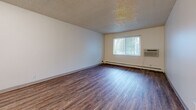 Located at 930-940 N Murray Blvd in Colorado Springs, Colorado, this one-bedroom, one-bathroom unit offers a practical and efficient layout across a single floor. The living room features rich, dark hardwood-style flooring, a textured ceiling, and a window with vertical blinds that brings in natural light. A wall-mounted air conditioning unit and baseboard heating provide year-round climate control. The kitchen is open and functional, outfitted with white cabinetry, white appliances including a range and refrigerator, and light wood-style flooring that ties the space together. A hallway connects the main living areas to the private bedroom, which is finished with plush carpet, a large window with vertical blinds, and a sliding-door closet offering ample storage. An additional closet provides further storage convenience. The bathroom features a full tub and shower combination surrounded by warm beige tile walls accented with a decorative mosaic tile border, chrome fixtures, and a clean, classic finish. The textured ceilings are consistent throughout, giving the unit a cohesive feel. The overall layout is compact yet well-organized, making efficient use of every room.