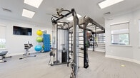 Located at 920 Yorkmont Ridge Lane in Fort Mill, South Carolina, this single-level fitness center is thoughtfully designed to support a wide range of workout styles and fitness goals. The space features a clean, modern aesthetic defined by light grey wood-look flooring, crisp white walls, crown molding, and bright panel lighting complemented by skylights that flood the interior with natural light.

The main workout area is anchored by a substantial multi-functional cable trainer and an expansive wall-mounted rig system equipped with TRX suspension trainers and monkey bars, enabling strength training, functional movements, and bodyweight exercises. A dedicated free weights section includes a full dumbbell rack, adjustable benches, and a barbell station, offering a complete strength training setup.

A separate functional training zone features a heavy punching bag suspended from an overhead rig, along with wall-mounted racks stocked with kettlebells, medicine balls, and slam balls—ideal for high-intensity and combat-style workouts. Additional equipment includes an ab bench, stretch equipment, and a collection of stability and exercise balls displayed on a rolling rack.

Windows along multiple walls invite natural light and outdoor views, while ceiling fans maintain airflow throughout. A water station and wall-mounted television are also present. The glass double-door entrance provides a welcoming, accessible entry point to this comprehensively outfitted facility.