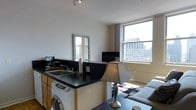 This single-floor urban studio offers an efficient and thoughtfully arranged open-plan layout that seamlessly integrates the living and kitchen areas. The kitchenette is equipped with dark countertops, a built-in sink, a coffee maker, and a washer unit conveniently tucked beneath the counter, maximizing functionality within the space. A decorative mirror with an ornate frame is mounted above the kitchen area, adding a stylish touch and reflecting natural light throughout. The living area features a comfortable sofa, a patterned area rug, and a coffee table, creating a cozy and welcoming atmosphere. A flat-screen TV is positioned to serve both the kitchen and living zones, enhancing the practical use of the open floor plan. Track lighting on the ceiling provides focused illumination, complementing the abundant natural light that streams through large windows. These windows are a standout feature, offering impressive elevated views of the surrounding urban skyline, with neighboring mid-rise buildings visible in the backdrop. The neutral white walls and clean lines throughout contribute to a bright, modern aesthetic. The space is well-suited for urban living, balancing comfort and practicality in a compact, light-filled setting.