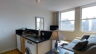 This single-floor urban studio offers an efficient and thoughtfully arranged open-plan layout that seamlessly integrates the living and kitchen areas. The kitchenette is equipped with dark countertops, a built-in sink, a coffee maker, and a washer unit conveniently tucked beneath the counter, maximizing functionality within the space. A decorative mirror with an ornate frame is mounted above the kitchen area, adding a stylish touch and reflecting natural light throughout. The living area features a comfortable sofa, a patterned area rug, and a coffee table, creating a cozy and welcoming atmosphere. A flat-screen TV is positioned to serve both the kitchen and living zones, enhancing the practical use of the open floor plan. Track lighting on the ceiling provides focused illumination, complementing the abundant natural light that streams through large windows. These windows are a standout feature, offering impressive elevated views of the surrounding urban skyline, with neighboring mid-rise buildings visible in the backdrop. The neutral white walls and clean lines throughout contribute to a bright, modern aesthetic. The space is well-suited for urban living, balancing comfort and practicality in a compact, light-filled setting.
