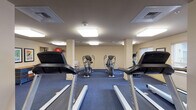 Located at 3500 S Sherman St in Englewood, Colorado, this single-floor fitness center is a well-equipped exercise facility designed to support a comprehensive workout routine. The space features neutral beige walls, durable blue carpet flooring, and flush overhead lighting panels that provide consistent, bright illumination throughout.

The cardio zone is prominently featured, housing a row of Spirit-brand treadmills positioned for comfortable use, accompanied by multiple elliptical cross-trainers and a recumbent bike. A multi-function cable and strength machine is also available for resistance training, offering users a variety of exercise options without the need for free weights.

A dedicated open floor area lined with large wall-to-wall mirrors serves as a stretching and functional fitness zone, giving users ample room for floor exercises, yoga, or cool-down routines. A mounted flat-screen television is positioned for easy viewing from the cardio equipment. Framed artwork adds a subtle decorative touch to the walls.

Natural light filters into portions of the space through windows fitted with horizontal blinds. A wall clock, towel storage cart, and exercise balls round out the practical amenities. The layout is thoughtfully organized, allowing users to move between equipment areas with ease, making this fitness center a functional and accessible resource for daily exercise.