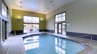This indoor pool area is designed to offer a comfortable and well-lit aquatic experience. The space features soaring ceilings that lend an open, airy feel, while large floor-to-ceiling windows along one wall flood the interior with natural light and provide views of an adjacent outdoor patio area, complete with seating. A set of glass-paneled doors provides direct access to the exterior, seamlessly connecting the indoor and outdoor environments.

The pool itself is generously sized, with calm, clear water and a tiled surround offering ample space for entry and movement along its edges. The interior walls are finished in a combination of neutral tones and an accent of olive green, adding a modern and refreshing aesthetic. Dark tile wainscoting runs along the lower portion of the walls, providing a clean visual boundary and a practical, moisture-resistant surface.

The overall ambiance is calm and functional, with recessed lighting supplementing the abundant natural light. The layout and design suggest a well-maintained, amenity-focused facility, suitable for both leisurely swimming and relaxation.