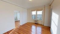 This single-floor space showcases timeless architectural character through its warm hardwood floors, which catch and reflect natural sunlight throughout the interior. The main room is generously proportioned, with white-painted walls and crown molding details that lend a classic, refined aesthetic. A pair of multi-pane windows line one wall, allowing abundant natural light to fill the space while offering views of the surrounding landscape, including what appears to be a scenic treeline in the distance. Beneath the windows sits a traditional cast-iron radiator, a hallmark of classic building construction that adds to the vintage character of the space. The ceiling is smooth and white, complementing the bright, airy atmosphere of the room. An open doorway leads directly into a bathroom, which is visible from the main space and features white fixtures, maintaining the clean and consistent color palette found throughout. The hardwood flooring extends seamlessly between rooms, reinforcing a sense of cohesion. Overall, the space blends period-appropriate architectural details with functional layout, offering a bright and welcoming environment with notable classic charm.