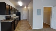 This single-floor residential space offers a practical and well-organized layout designed for comfortable everyday living. The kitchen is outfitted with dark espresso cabinetry that provides ample storage, complemented by granite-look countertops, a double basin sink, and black appliances including a refrigerator, stove, and overhead microwave. The kitchen flows directly into a central hallway, creating an open and connected feel throughout the unit. Hardwood-style flooring runs through the kitchen and hallway, adding warmth and visual continuity, while carpeting transitions into additional living areas. The walls are finished in a clean, neutral light gray tone that brightens the interior and serves as a versatile backdrop. The hallway leads to interior doors, offering clear separation between living and private areas. An electrical panel is conveniently accessible along the hallway wall. Flush-mount ceiling lighting provides consistent illumination throughout. The overall ambiance is clean, modern, and move-in ready, with a straightforward layout that maximizes the available space on a single level.