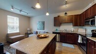 This single-floor residence offers a thoughtfully designed open-concept layout that seamlessly connects the kitchen, dining, and living areas. The kitchen serves as a focal point, featuring rich dark-wood cabinetry extending to the ceiling, granite countertops, a full suite of stainless steel appliances including a refrigerator, range, microwave, and dishwasher, and a generously sized island with pendant lighting overhead. Track lighting throughout the kitchen and living area provides flexible illumination. Wood-look flooring flows through the main living spaces, contributing to a warm, cohesive aesthetic.

The dining area benefits from direct access to the outdoors through a glass door framed by large windows that bring in ample natural light. The bedroom features warm-toned accent walls, plush carpeting, and a window with blinds offering privacy and natural light. A hallway connects the bedroom to the full bathroom, which includes a dual-sink vanity with granite counters, dark wood cabinetry, a large mirror, and a tiled tub-and-shower combination. The overall design maintains a consistent palette of warm neutrals and dark wood finishes throughout, creating a unified and comfortable living environment.