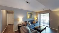 This single-floor space offers a comfortable and thoughtfully designed living environment. The living room serves as the centerpiece, finished in soft blue-grey tones that create a calm and inviting atmosphere. The room is furnished with plush blue-grey sofas accented with patterned and yellow throw pillows, adding a touch of warmth and character to the space. A dark wood coffee table anchors the seating area, complementing the rich hardwood flooring that runs throughout. A decoratively framed mirror with a gold finish is mounted on the accent wall, enhancing the sense of depth and light in the room. Ambient lighting is provided by matching modern table lamps with warm-toned shades, contributing to a cozy, welcoming glow. A large sliding glass door along one wall floods the space with natural light and offers views of the greenery outside, seamlessly connecting the interior to the outdoors. Ceiling vents are discreetly integrated into the white ceiling, maintaining a clean, uncluttered aesthetic. The overall ambiance is one of relaxed sophistication, blending neutral architectural finishes with carefully chosen furnishings to create a comfortable and functional living environment.