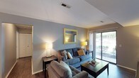 This single-floor space offers a comfortable and thoughtfully designed living environment. The living room serves as the centerpiece, finished in soft blue-grey tones that create a calm and inviting atmosphere. The room is furnished with plush blue-grey sofas accented with patterned and yellow throw pillows, adding a touch of warmth and character to the space. A dark wood coffee table anchors the seating area, complementing the rich hardwood flooring that runs throughout. A decoratively framed mirror with a gold finish is mounted on the accent wall, enhancing the sense of depth and light in the room. Ambient lighting is provided by matching modern table lamps with warm-toned shades, contributing to a cozy, welcoming glow. A large sliding glass door along one wall floods the space with natural light and offers views of the greenery outside, seamlessly connecting the interior to the outdoors. Ceiling vents are discreetly integrated into the white ceiling, maintaining a clean, uncluttered aesthetic. The overall ambiance is one of relaxed sophistication, blending neutral architectural finishes with carefully chosen furnishings to create a comfortable and functional living environment.