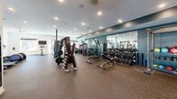 Located at 2418 Main St in Rocky Hill, CT, this well-appointed fitness center offers a comprehensive exercise environment across a single, generously proportioned floor. The space is thoughtfully designed with durable speckled rubber flooring throughout, providing both safety and comfort underfoot during workouts.

The main exercise area is organized into distinct workout zones. The cardio section features multiple treadmills, elliptical machines, and a stationary bike, all positioned near windows with roller shades that allow natural light to filter in. A Peloton cycling bike and an interactive digital fitness display add modern, tech-forward options for guided workouts.

The strength training area is lined with a deep teal accent wall, creating a visually distinct and energizing atmosphere. A full rack of dumbbells in graduated weights, multiple cable and resistance machines, and free weight benches are neatly arranged alongside floor-to-ceiling mirrors that enhance the sense of space. Athletic artwork on the walls contributes to the motivating ambiance.

Additional amenities include a water fountain, recycling stations, ceiling fans for climate comfort, and recessed lighting throughout. A separate room off the main floor, visible through glass doors, offers additional utility space. The overall design is clean, modern, and functional, making this fitness center a well-equipped destination for a wide range of training needs.