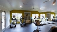 This fitness center is a single-floor space designed to support a well-rounded cardio workout experience. The room is immediately distinguished by its bold yellow walls, which create an energizing and motivating atmosphere for exercise. Large, multi-pane windows line the walls on multiple sides, flooding the space with abundant natural light and offering views of the surrounding greenery outdoors. The vaulted ceiling adds a sense of openness and volume to the room, complemented by a ceiling fan that aids in air circulation during workouts. Recessed lighting provides additional illumination throughout. The facility is outfitted with a variety of cardio equipment, including treadmills, elliptical machines, and a stationary bike, all arranged to allow comfortable use and movement between machines. A built-in shelving unit at the center of the room provides organized storage, and a water cooler is conveniently positioned nearby. The dark rubberized flooring is both practical and durable, suitable for high-traffic fitness use. Overall, the space strikes a balance between functionality and an inviting aesthetic, making it a well-appointed facility for daily exercise.