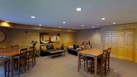 This single-floor space functions as a comfortable lounge and gathering area, designed with a warm and welcoming ambiance. The room features earthy, golden-toned walls accented by a textured ceiling with recessed lighting, creating a well-lit yet cozy atmosphere throughout.

The seating area is anchored by two dark leather sofas arranged around a wooden coffee table, offering a relaxed setting for conversation or lounging. Decorative throw pillows and a bold piece of abstract wall art add visual interest and warmth to this section of the room.

Adjacent to the lounge area is a dining space furnished with a sturdy wooden high-top table surrounded by matching bar-height chairs, making it suitable for casual dining or group gatherings. Additional side seating and small accent tables are positioned near the walls, maximizing the use of the available space.

The carpeted flooring adds comfort underfoot and complements the overall neutral color palette of the room. A doorway on the far wall provides access to an adjoining area. The combination of functional furniture arrangements and thoughtful décor creates a versatile, multi-purpose space suited for both socializing and dining.