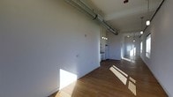 This single-floor space showcases a compelling blend of industrial and modern design. The open-concept layout is anchored by rich hardwood flooring that stretches throughout, reflecting abundant natural light that pours in and creates striking geometric patterns across the floor. Soaring ceilings draw the eye upward to exposed ductwork — a defining industrial design feature — that runs the length of the space, adding character and an urban loft aesthetic. Pendant lighting fixtures hang at varying heights, providing focused illumination while complementing the raw, contemporary style. The kitchen area is positioned toward the rear of the main space and features white appliances, maintaining a clean and functional look. Crisp white walls offer a neutral backdrop that enhances the brightness and openness of the interior. The layout flows naturally from the main living area through to additional rooms, connected by an open corridor that maintains the airy, unobstructed feel of the space. Overall, the combination of exposed mechanical elements, warm wood flooring, and generous natural light creates an inviting yet architecturally distinctive environment well-suited for a variety of uses.