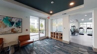 Located at 2429 Americana Blvd in Orlando, Florida, this fitness studio is a thoughtfully designed, single-floor facility offering a comprehensive workout environment. The space is accessed through a stylish lounge-style entry area featuring patterned carpet flooring, a bold dark teal ceiling, a wood cubby storage unit, and an eye-catching typographic glass partition accented with motivational words. Comfortable leather seating and large glass doors fill the entryway with natural light and views of the surrounding landscaped exterior.

The main gym floor transitions to durable rubber flooring and is bathed in natural light through expansive floor-to-ceiling windows overlooking lush outdoor greenery. The ceiling features decorative wood slat accents alongside recessed lighting, lending a contemporary, upscale aesthetic. The facility is equipped with multiple treadmills, stair climbers, and a variety of strength-training machines, including cable and resistance equipment. A dedicated functional training zone offers TRX suspension systems, squat racks, and open floor space, while a mirrored stretching and free-weight area with a decorative lattice wall accent rounds out the offerings. Together, these zones provide a well-rounded fitness experience suited to a wide range of training styles and fitness levels.