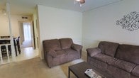 This single-story space includes a living area finished with wall-to-wall carpeting in a warm beige tone, contributing to a casual and comfortable atmosphere. The walls are painted in a light, neutral hue, keeping the space feeling open and understated. A ceiling light fixture provides overhead illumination for the room. The living area accommodates a brown microfiber loveseat and matching armchair arranged around a dark-framed coffee table, creating a functional seating arrangement. A doorway visible to the left side of the living area suggests a connected hallway that leads to additional rooms, hinting at a straightforward, practical layout throughout the home. The overall aesthetic is modest and utilitarian, with minimal decorative elements, making the space well-suited for everyday living. The single-floor layout offers convenient accessibility throughout.