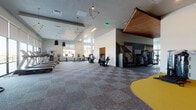 This expansive fitness center is thoughtfully organized into several distinct workout zones, each designed to accommodate a comprehensive range of exercise disciplines. The space is characterized by soaring ceilings, large floor-to-ceiling windows that flood the interior with natural light, and views of the surrounding exterior. Patterned carpet tile flooring covers the main areas, while select zones feature specialized flooring suited to their intended use.

The cardio zone is lined with a row of treadmills positioned directly in front of expansive windows, offering open views during workouts. Nearby, upright bikes, stair climbers, and elliptical machines are arranged across a generously sized open floor. A dedicated strength training area houses power racks, barbells, weight plates, dumbbells on tiered racks, and a variety of cable and selectorized machines, some positioned on bold yellow accent flooring. Full-length mirrors throughout this zone enhance both functionality and the sense of depth.

A separate studio room features warm wood-tone flooring, mirrored walls, decorative pendant lighting, and space for cycling classes and functional training. Geometric and globe-style pendant fixtures add a modern, design-forward touch throughout, complemented by a striking wood-slat ceiling accent that grounds the overall aesthetic.