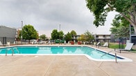 Located at 1117 City Park Ave in Fort Collins, Colorado, this outdoor amenity space centers around a generously sized rectangular swimming pool with clear, blue water and classic tiled edges. The expansive concrete pool deck surrounds the pool on all sides, providing ample space for relaxation and recreation. Numerous lounge chairs and sun loungers are arranged around the perimeter, offering plenty of seating for residents to enjoy the outdoors. Vibrant red seating accents add a pop of color against the neutral tones of the deck and the dark-painted building facades nearby. The area is enclosed by a sleek, black iron fence, providing both security and an open, airy feel. Tall, mature trees are positioned throughout and around the space, offering natural shade and a lush, park-like atmosphere. Decorative white planters with flowering plants add charming touches of greenery at the fence line. The surrounding multi-story residential buildings, finished in dark gray and muted tones, frame the space and create a cohesive community environment. The well-maintained lawn areas adjacent to the pool contribute to the overall inviting and resort-like character of this outdoor gathering space.