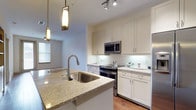 This single-floor residence offers a thoughtfully designed living space that balances modern aesthetics with practical functionality. The open-concept kitchen serves as a central highlight, showcasing granite countertops with an undermount stainless steel sink set into a generous island that doubles as a breakfast bar. Crisp white shaker-style cabinetry lines the walls, providing ample storage while contributing to the bright, clean atmosphere of the space. Stainless steel appliances, including a French door refrigerator, electric range, and over-the-range microwave, lend a contemporary, professional feel to the kitchen. Track lighting on the ceiling works in tandem with elegant pendant lights hanging above the island, ensuring the workspace is well-lit and visually inviting. The warm hardwood flooring adds a natural contrast to the light cabinetry and countertops, creating a cohesive and welcoming ambiance. Natural light filters in through nearby windows, enhancing the airy, open feel of the space. The layout flows seamlessly from the kitchen toward the living area, making the home well-suited for both everyday living and entertaining. Overall, the space presents a clean, modern design with quality finishes throughout.