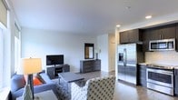 This single-floor space showcases a thoughtfully designed open-concept layout, where the living area flows naturally into the kitchen. The living room is furnished with a plush sofa, a patterned accent chair, and a sleek coffee table, all arranged around a flat-screen television mounted atop a low-profile media console. A framed mirror and a matching dark-toned dresser add depth and dimension to the space, complementing the light hardwood-style flooring that runs throughout.

The kitchen is partially visible and features dark upper cabinetry paired with a stainless steel French door refrigerator, lending a polished, modern aesthetic. The transition between living and kitchen areas is defined by a subtle change in flooring, maintaining a cohesive yet distinct separation between the two zones.

The walls are finished in crisp white, contributing to a bright and airy atmosphere, while recessed lighting ensures consistent illumination throughout. The overall design palette relies on a contrast of dark and light tones, creating a contemporary and clean ambiance well-suited for everyday living.