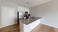 This single-floor unit showcases a clean, contemporary design centered around an open-concept layout that seamlessly connects the kitchen and living area. The living space is bright and airy, featuring light wood-tone laminate flooring, neutral gray walls, and recessed lighting throughout. A large sliding glass door fitted with vertical blinds allows natural light to fill the room while providing access to the outdoors.

The kitchen is well-appointed with white shaker-style cabinetry extending to the ceiling for ample storage, complemented by a dark gray geometric tile backsplash. Stainless steel appliances include a full-size refrigerator, electric range, and built-in microwave. A spacious peninsula-style island with a gray quartz countertop features an undermount double sink and additional cabinetry below with brushed hardware, offering both prep space and a natural divide between the kitchen and living area.

The bathroom includes a full tub-shower combination with clean white surrounds, a white vanity with dark countertop, and dark wood-tone flooring that adds contrast to the space. Overall, the unit presents a cohesive, modern aesthetic with functional finishes suited for everyday living.
