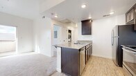 This single-floor, one-bedroom, one-bathroom unit offers a well-organized layout across a compact but functional footprint. The combined kitchen and living area serves as the heart of the space, where the kitchen is equipped with dark espresso cabinetry, granite-look countertops, stainless steel appliances including a gas range, microwave, refrigerator, and dishwasher. A peninsula with seating overhang provides both additional prep space and a casual dining option. Light wood-look flooring runs throughout the kitchen area, lending a clean, contemporary feel. The living area transitions to soft carpeting and features a sliding glass door that opens to a private balcony, allowing natural light to fill the room and offering outdoor views. A dedicated in-unit laundry area is conveniently accessible from the main living space. The bedroom is carpeted and bright with natural light, featuring a sliding-door closet with a built-in shelf and hanging rod. A hallway connects the bedroom to the full bathroom, which includes a tub-shower combination, wood-look tile flooring, and a vanity with dark cabinetry. The overall palette of white walls and neutral finishes creates a clean, cohesive ambiance throughout.