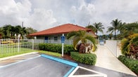 This single-story commercial building presents a clean, functional design well-suited to its tropical surroundings. The structure is immediately recognizable by its prominent red metal hip roof, which crowns light gray exterior walls and lends the building a distinctive, polished appearance. A bold blue entry door provides a striking contrast against the neutral facade, while a standard window allows natural light to filter into the interior space.

The building is fully accessible, as indicated by designated accessible parking spaces directly in front of the entrance, complete with proper signage and marked curb cuts. The parking area is paved and well-maintained, offering convenient on-site access for visitors and customers.

Thoughtfully maintained landscaping surrounds the structure, featuring lush tropical vegetation including mature palm trees, ornamental shrubs, and colorful foliage plants that enhance the building's curb appeal. A white perimeter fence adds definition to the property boundary while contributing to the overall neat, organized appearance of the site.

The building appears to be part of a larger facility, with additional structures visible in the background. The overall setting reflects a well-kept commercial property designed for practical use in a warm, tropical climate.